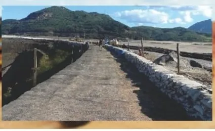 Pista sobre a crista de barragem, com enrocamento na lateral e montanhas ao fundo