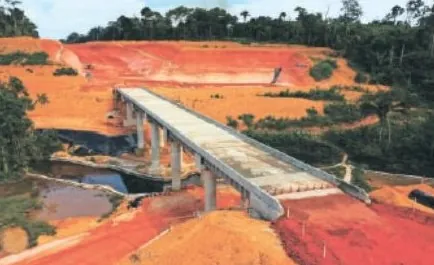 Ponte de concreto sobre curso d’água em estrada de terra vermelha, com floresta ao fundo
