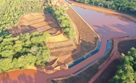 Vista aérea de reservatório de água avermelhada cercado por floresta, com canal revestido e taludes em bancadas