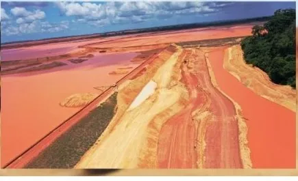 Vista aérea de berma em terra ao longo de reservatório de água avermelhada, com floresta à direita